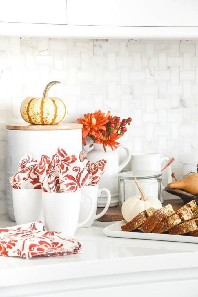 Fall coffee bar styled on a white kitchen counter with mugs, patterned napkins, small pumpkins, fall flowers, and a plate of sliced bread.