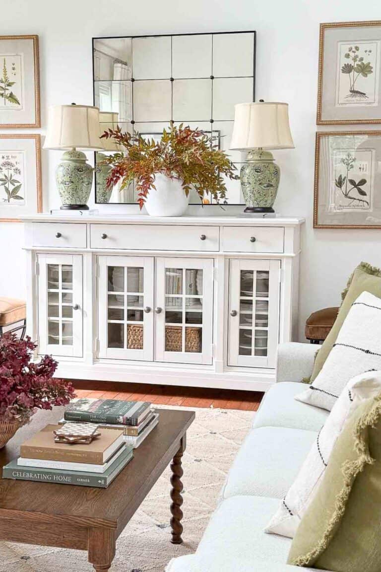Fall living room with a white buffet, a vase of colorful autumn branches, green table lamps, and cozy sofa pillows.