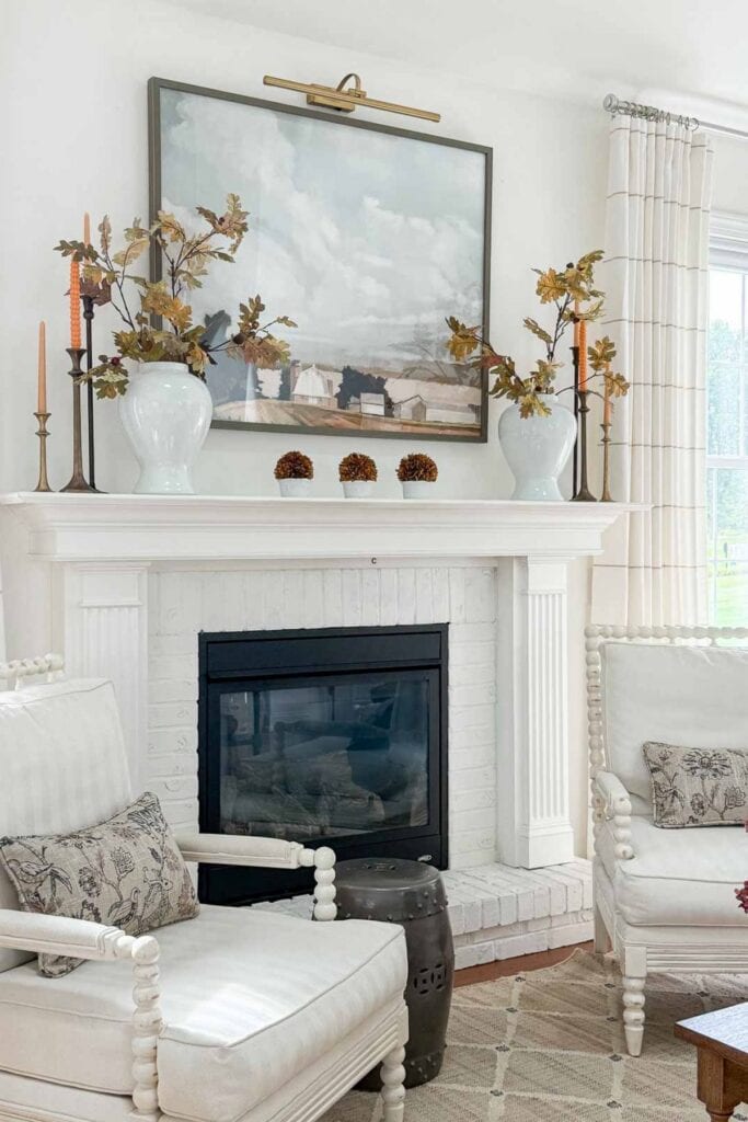 Fall mantel decorated with white vases of autumn branches, candlesticks, and a large framed painting above the fireplace.