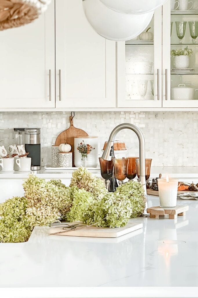 White kitchen with hydrangeas in the sink, amber glassware, a lit candle, and a fall coffee bar styled on the counter in the background.