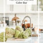 White kitchen decorated for fall with hydrangeas in the sink, amber glassware, a lit candle, and a fall coffee bar styled on the counter.