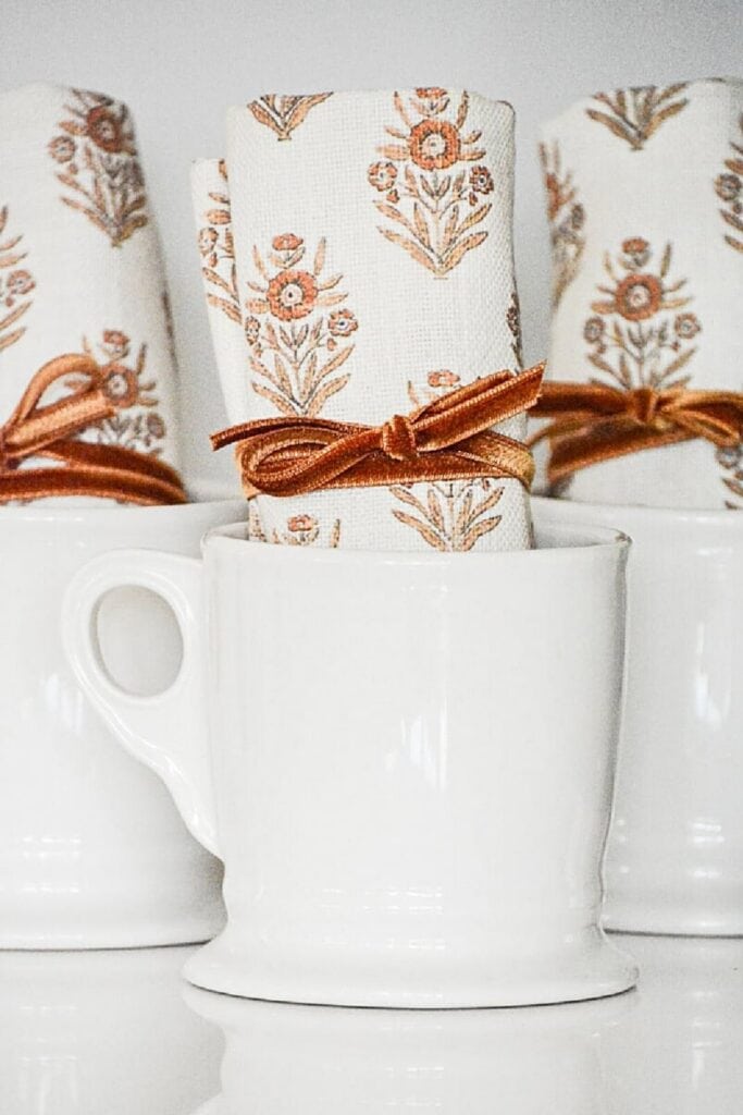 White mugs with fall patterned napkins tied with velvet ribbon, styled for a cozy fall tea bar.
