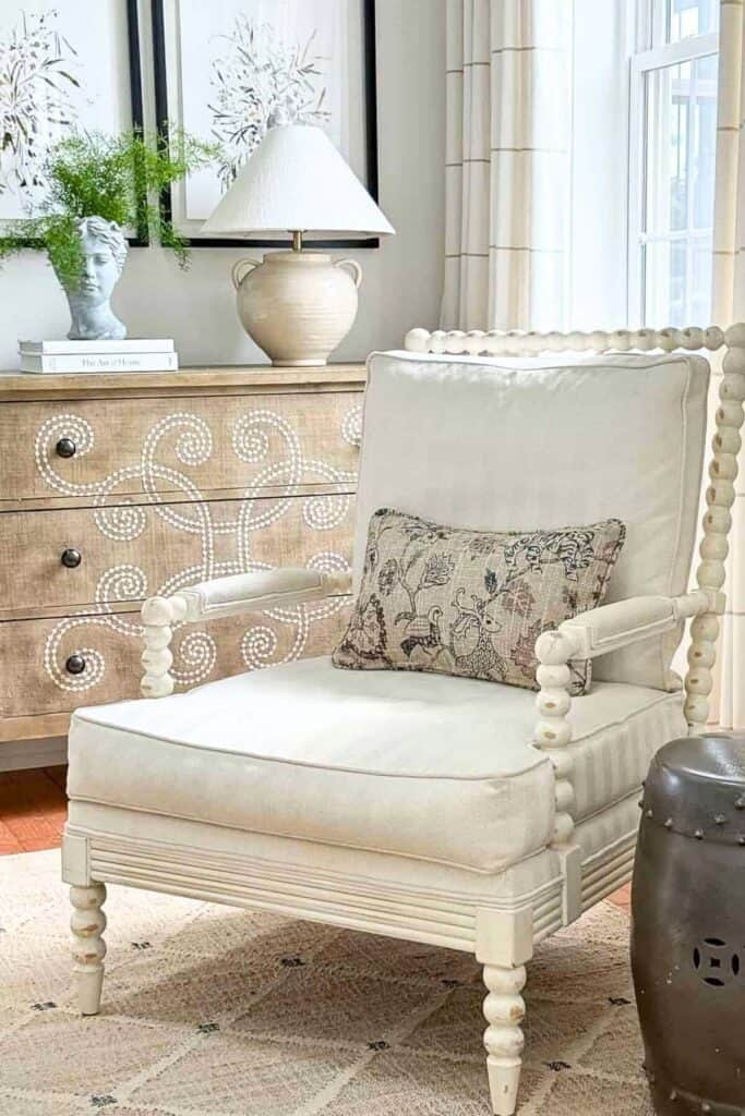 Neutral accent chair styled with a single patterned pillow beside a wood dresser and lamp for an uncluttered, calm living room look