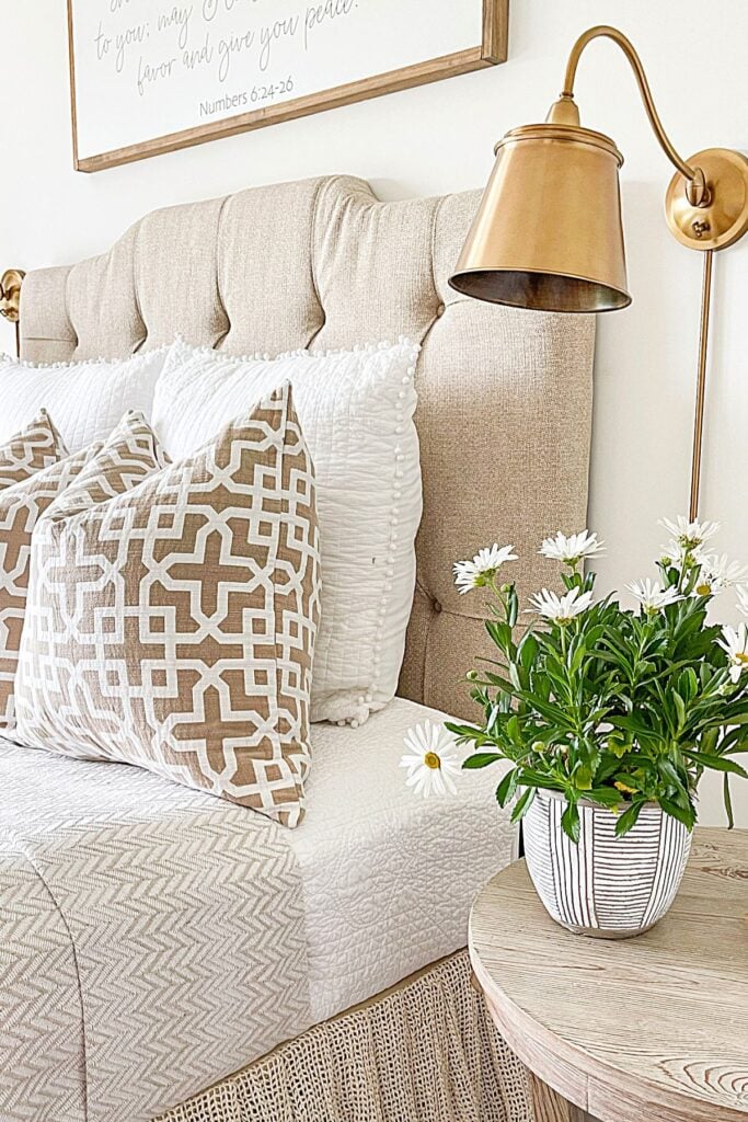 organized bedroom with neutral bedding, pillows, wall lamp, and simple nightstand styling with flowers