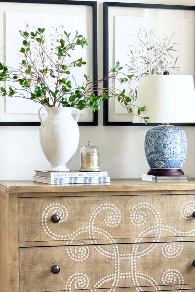 Console table styled with greenery in a white urn, stacked books, a blue lamp, and framed art for an uncluttered, balanced look