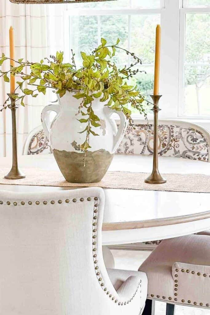 Simple dining table centerpiece with greenery in a white urn flanked by brass candlesticks creating an uncluttered, calm look