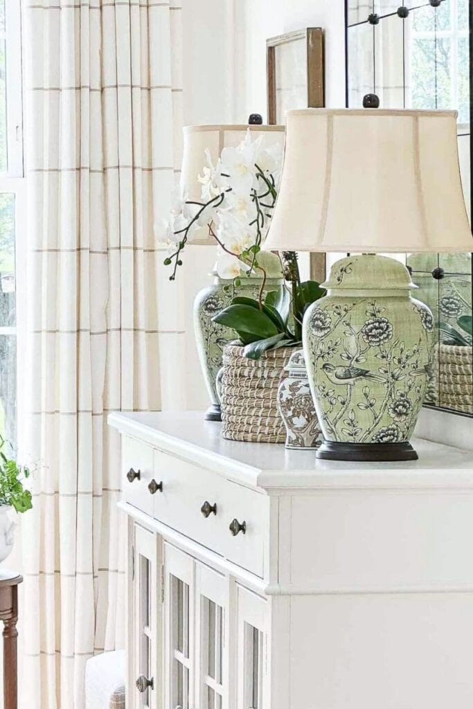 White buffet styled with green ceramic lamps, a basket with orchids, and a mirror for a calm, uncluttered look