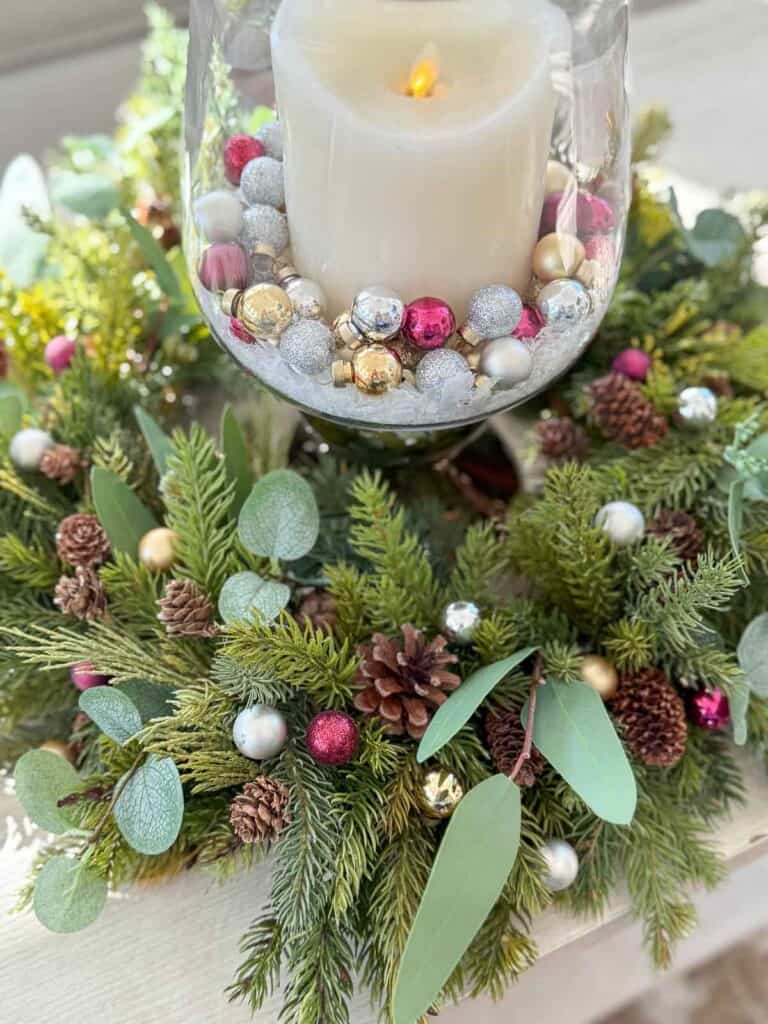 Christmas candle ring made with faux evergreens, eucalyptus, pinecones, and small colorful ornaments around a hurricane candle