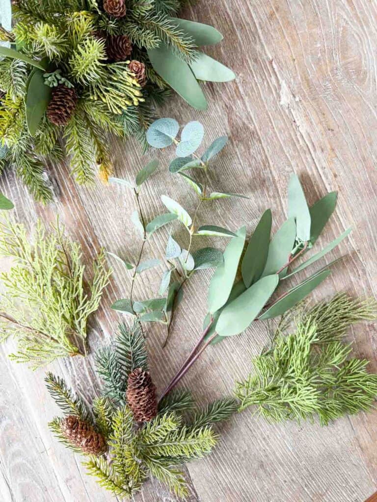 Faux evergreen branches, eucalyptus stems, and pinecones laid out on a table for a Christmas wreath DIY
