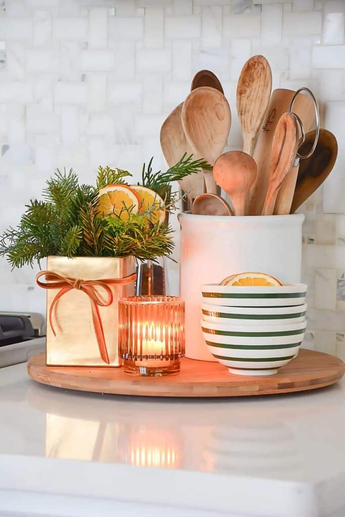 small arrangement of faux Christmas greens with dried oranges styled on a kitchen tray with a candle and wooden spoons