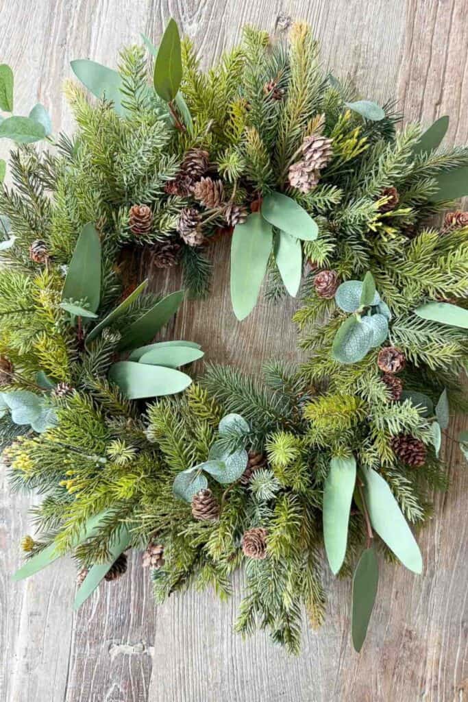 Christmas wreath made with faux evergreens, pinecones, and eucalyptus leaves laying on a wooden table