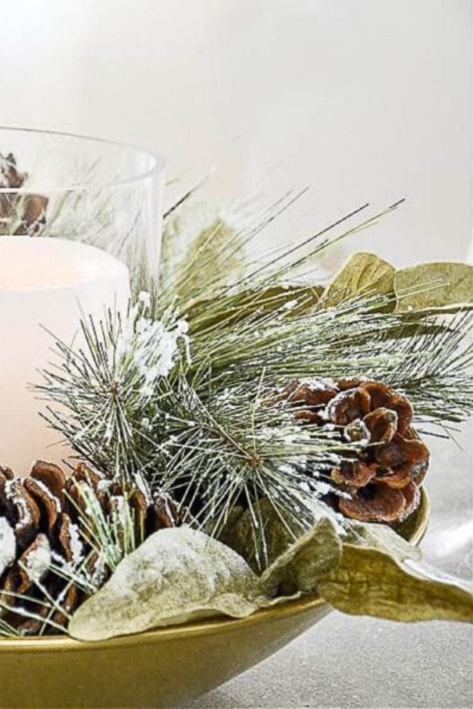 close up of faux Christmas greens and snowy pinecones surrounding a candle in a gold bowl