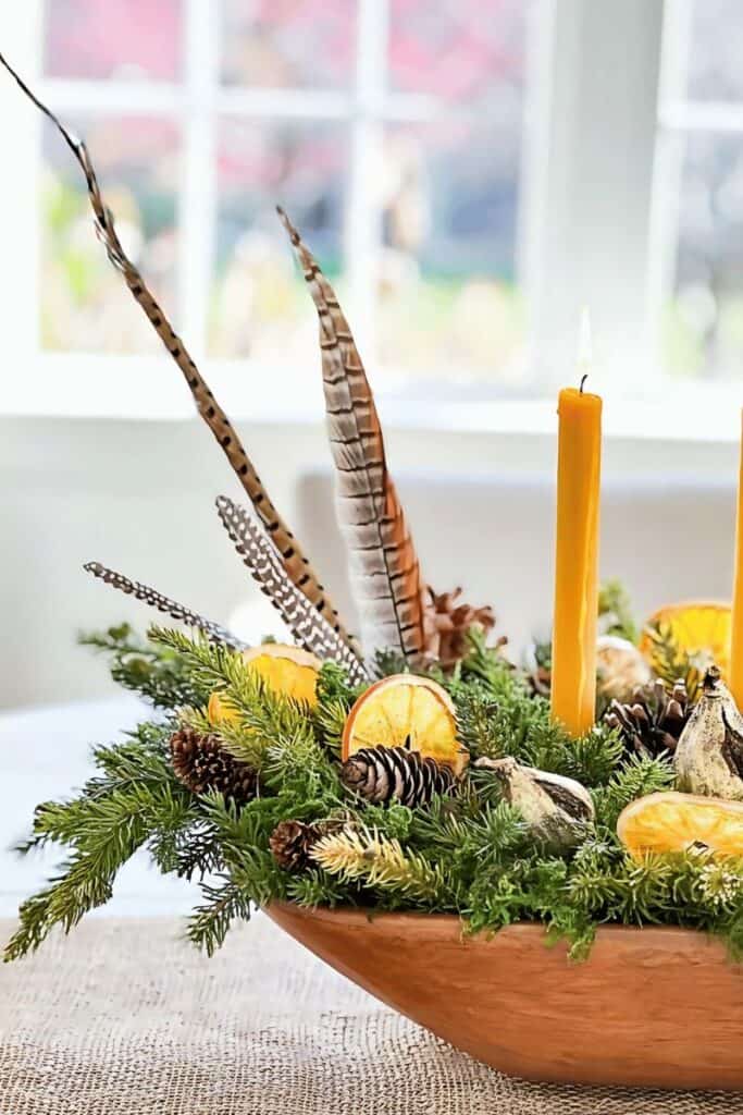 faux Christmas greens arranged in a wooden dough bowl with pinecones, candles, and pheasant feathers