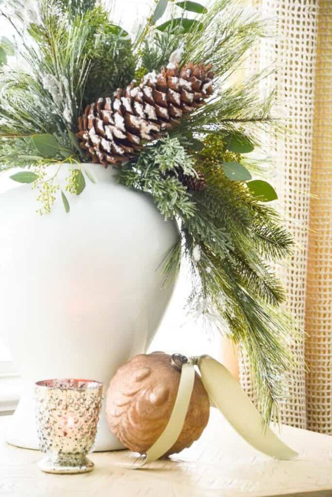 white vase filled with faux Christmas greens, eucalyptus, and snowy pinecones with a candle and ornament on the table