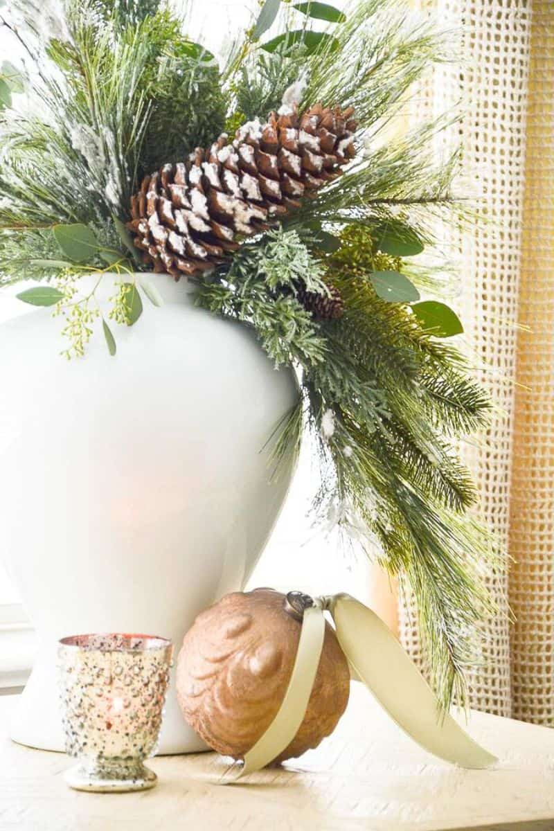 white vase filled with faux Christmas greens, eucalyptus, and snowy pinecones with a candle and ornament on the table