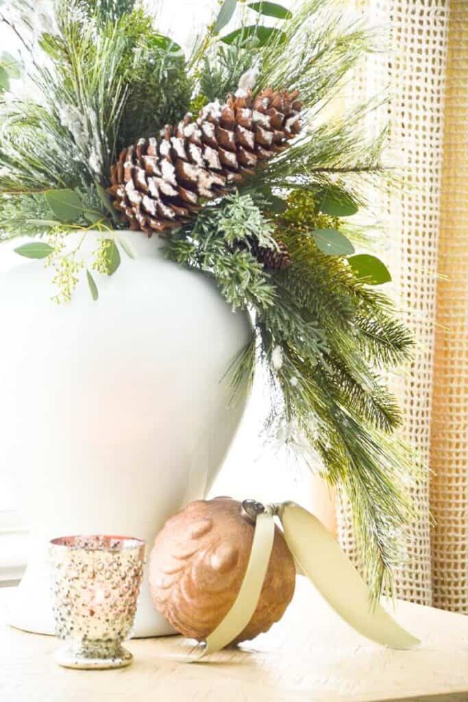 a 3-step Christmas arrangement made with a white vase, greens, and a large pine cone