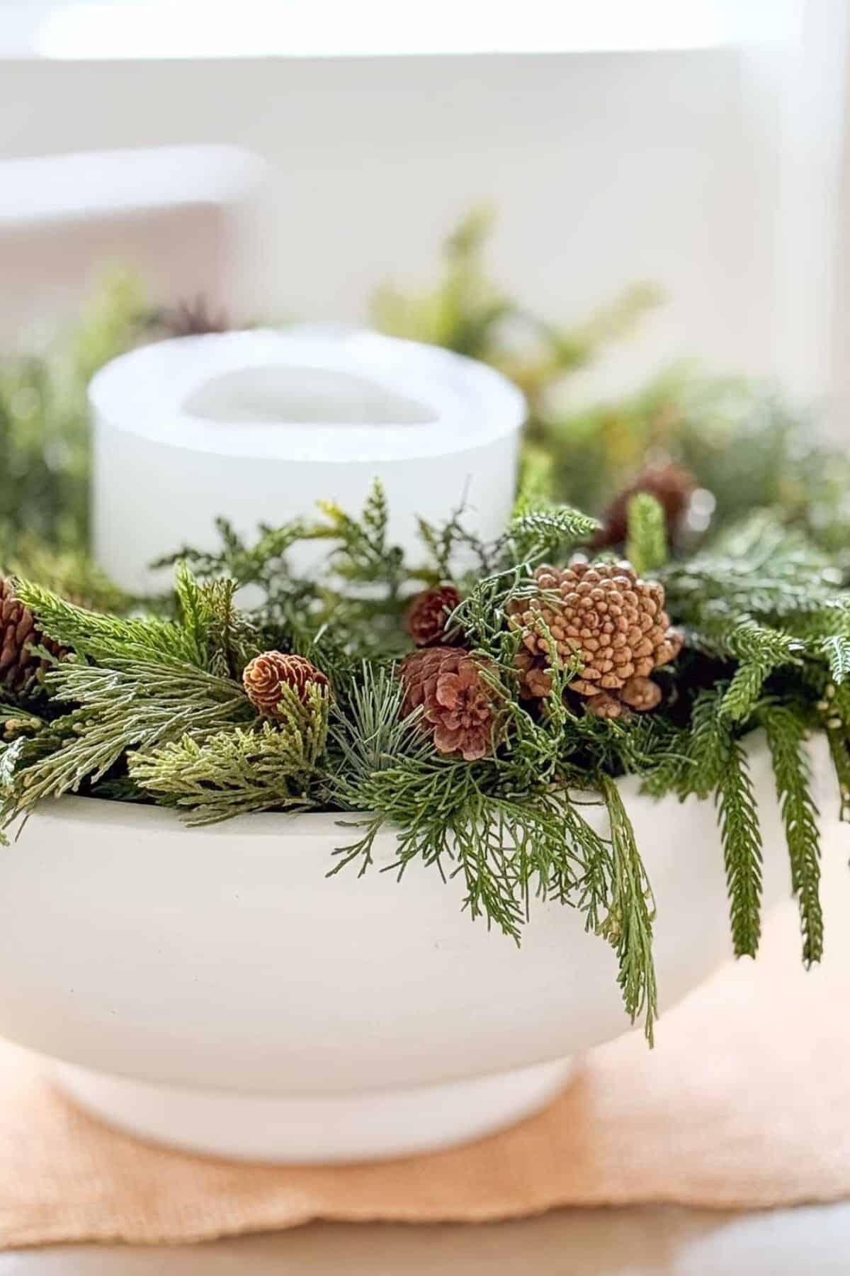 3- step Christmas arrangement- a white bowl filled with a variety of greens and pinecones and a candle