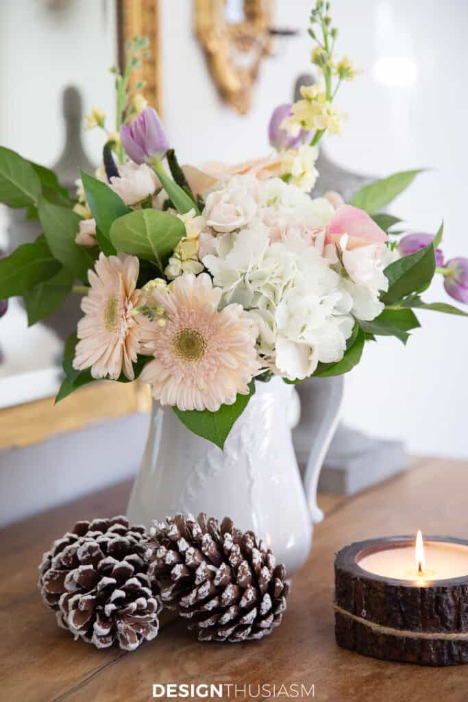 a vase of flowers, pinecones and a small candle. on a table