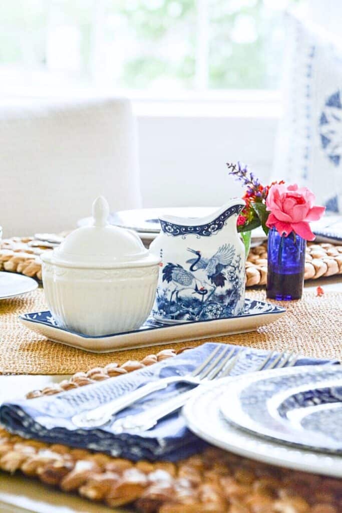 dining table set with a blue and white chinoiserie pitcher and white sugar bowl on a serving tray, a small cobalt blue vase with a pink rose and wildflowers, woven chargers, blue and white napkins, and layered china place settings