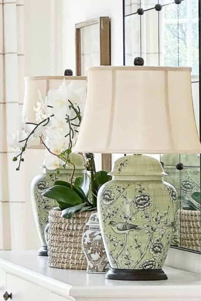 white buffet styled with a pair of celadon green chinoiserie ginger jar table lamps with cream shades, a white orchid plant in a wicker basket, and an antique mirrored cabinet in a neutral living room