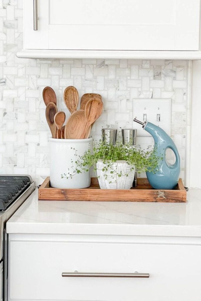 white kitchen counter styled with a wooden tray holding a white crock of wooden spoons, a small potted herb plant, and a blue ceramic olive oil dispenser on a marble tile backsplash