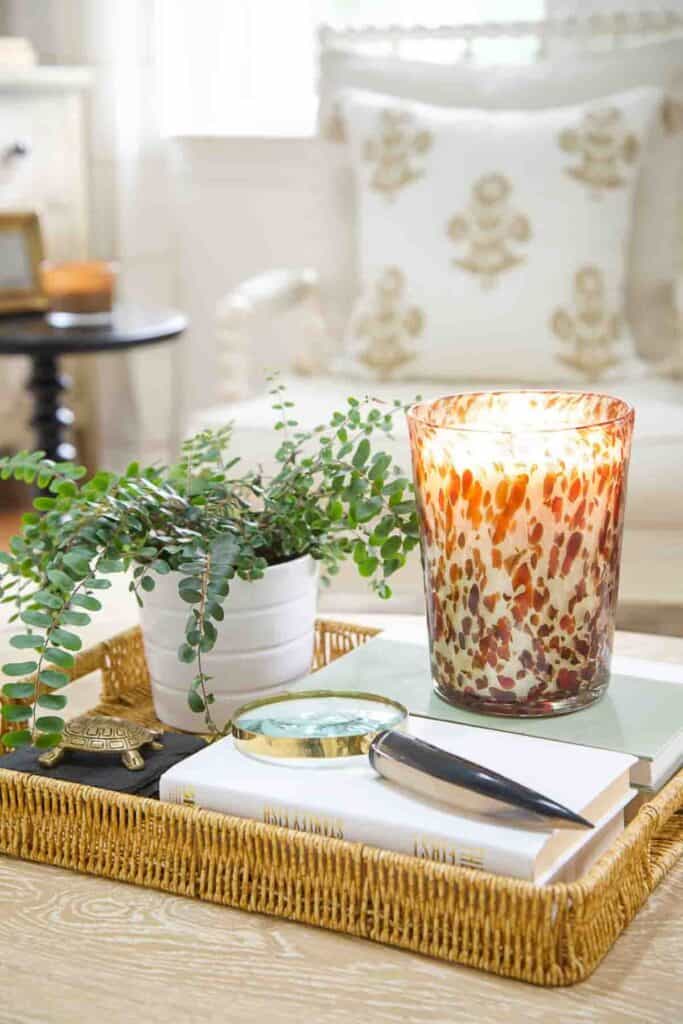 wicker tray on a coffee table styled with a glowing tortoiseshell glass candle, a white potted fern, stacked books, a brass magnifying glass, and a small brass turtle figurine in a neutral living room