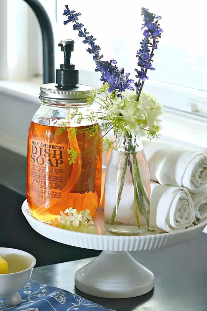 a pedestal cake plate with dish soap, bar towels and a vase of flowers on it
