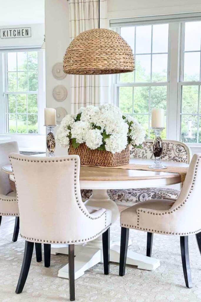ound dining room table styled with a wicker basket filled with white hydrangeas, crystal pillar candle holders, linen upholstered chairs with nailhead trim, and a woven pendant light in a bright neutral dining room