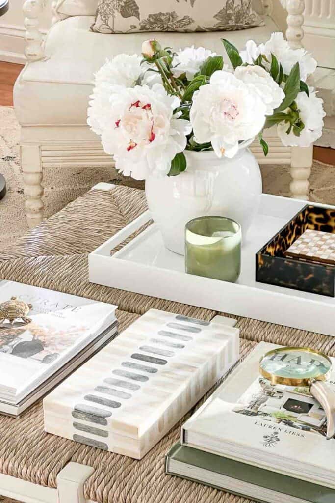 woven coffee table styled with a white lacquer tray holding a round white vase of white peonies and a green glass candle, a tortoiseshell box, stacked decorating books, a mother of pearl decorative box, and a brass magnifying glass in a neutral living room