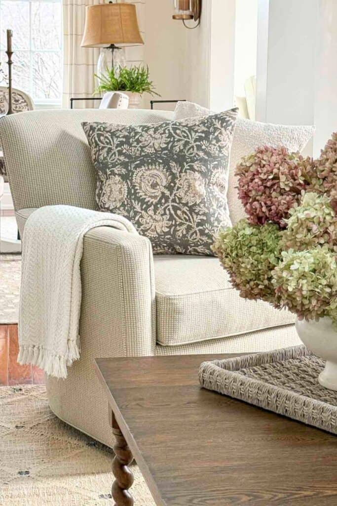 neutral linen armchair with a gray and cream floral pillow and a white fringed throw draped over the arm, beside a wood barley twist coffee table holding a woven tray and a white vase of dried pink and green hydrangeas in a bright neutral living room