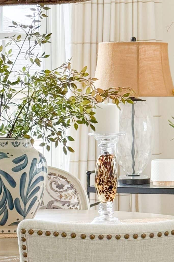 neutral dining room vignette with a large blue and white planter overflowing with green branches, a crystal pillar candle holder with a tortoiseshell glass insert, a glass table lamp with a linen shade, and a white essential oil diffuser on a black side table