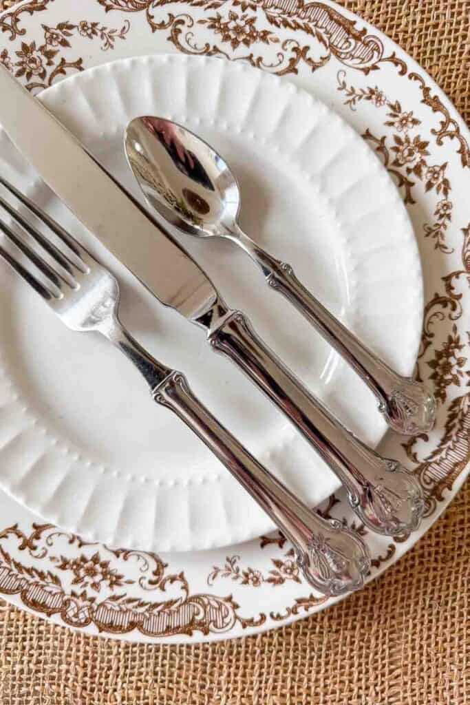 close up of a brown and white transferware dinner plate with polished silver flatware including a fork, knife, and spoon laid on top, resting on a woven wicker charger