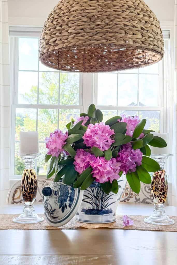 dining table centerpiece with a large blue and white chinoiserie planter overflowing with pink rhododendrons and green leaves, flanked by tortoiseshell crystal pillar candle holders on a jute table runner beneath a woven seagrass pendant light