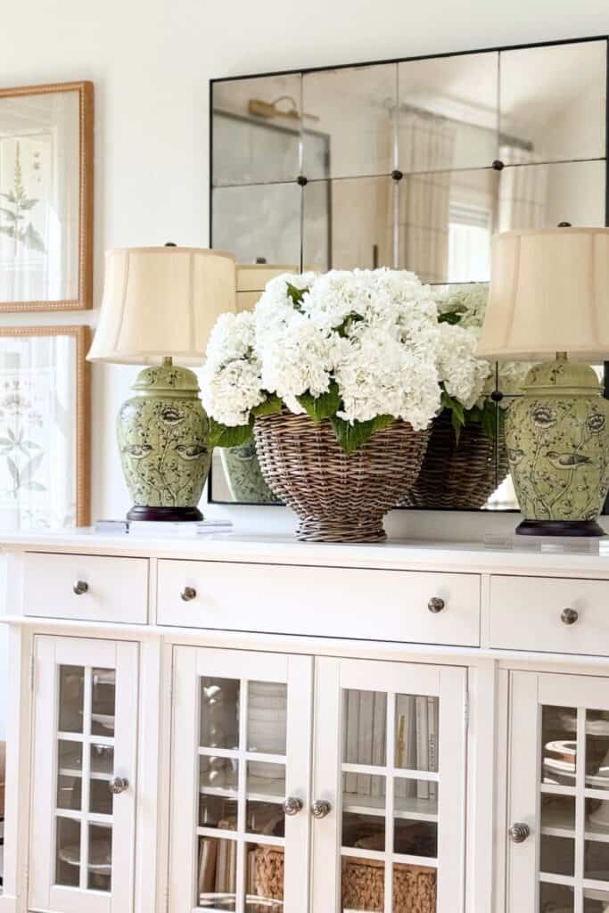 White hydrangea in a woven basket flanked by two green chinoiserie lamps on a white buffet