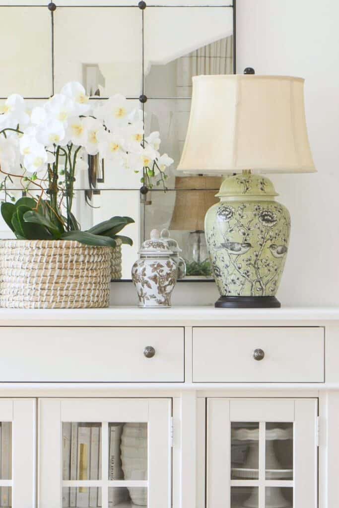 Anchor decor on a white buffet, a green chinoiserie lamp, white orchid in a woven basket, and a small ginger jar