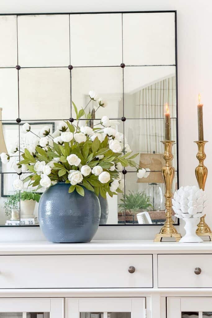 Lit brass candlesticks of varying heights with a white ceramic artichoke finial on a white buffet