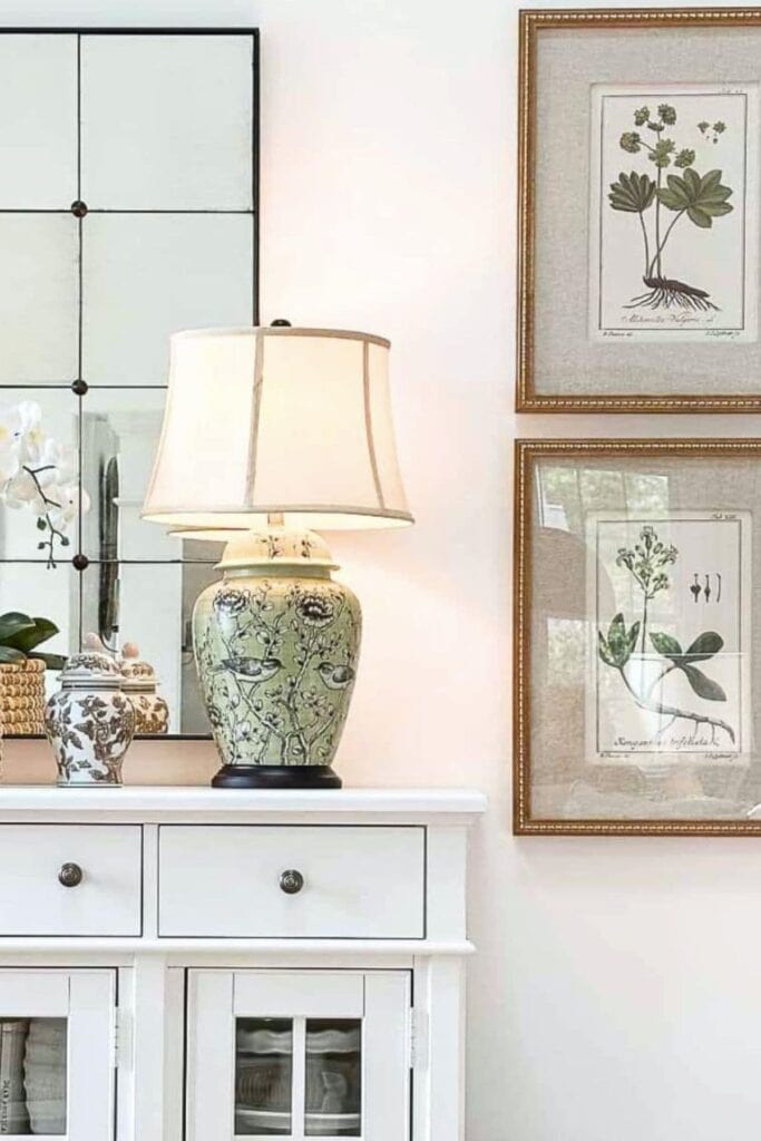 Lit green chinoiserie lamp on a white buffet with botanical art and ginger jars nearby