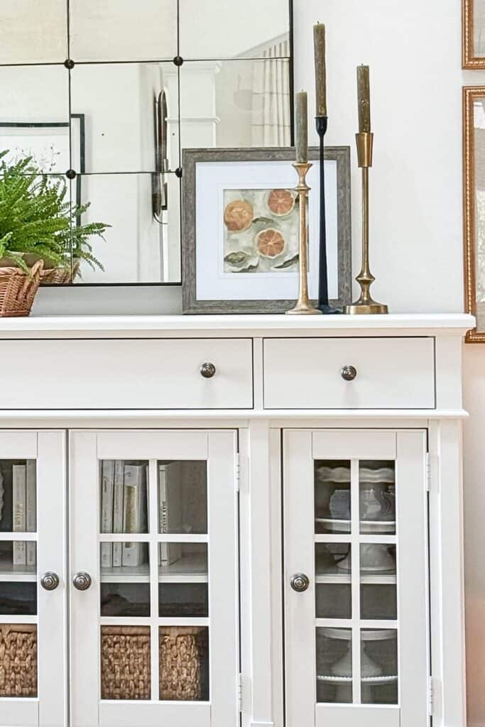 Framed citrus print leaned against a mirror on a white buffet with brass and black candlesticks of varying heights