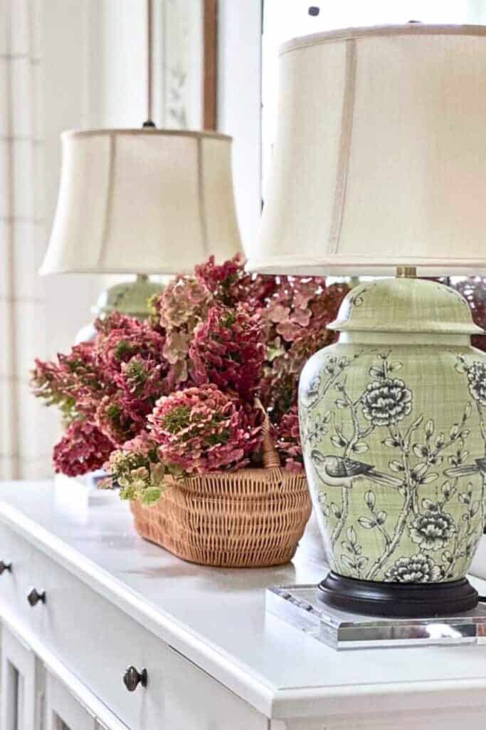 Dried burgundy hydrangea in a woven basket beside a green chinoiserie lamp on a white buffet