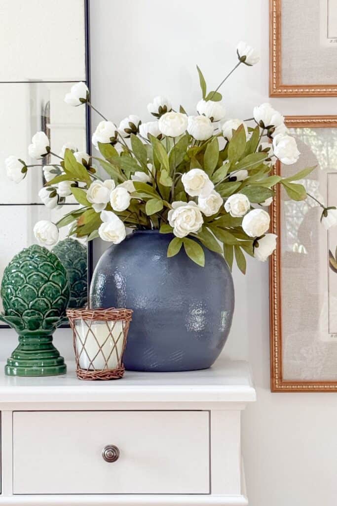 Grouping of three items on a white buffet, a navy vase with white florals, a green artichoke finial, and a woven candle