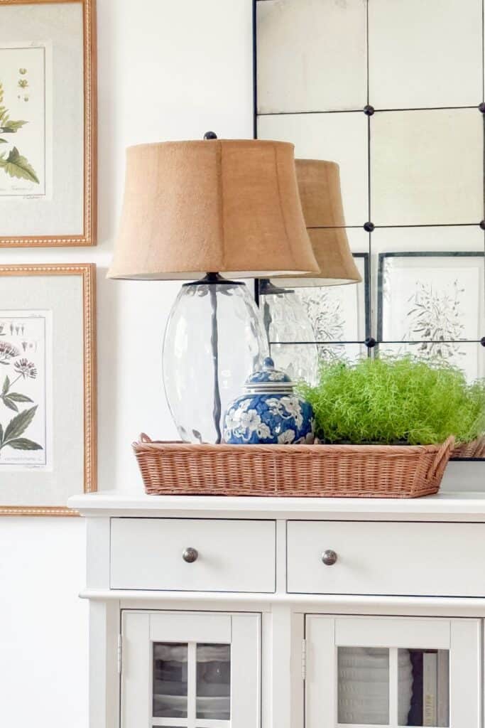 Buffet vignette showing mixed textures, a glass lamp, blue and white ginger jar, woven basket tray, and faux greenery