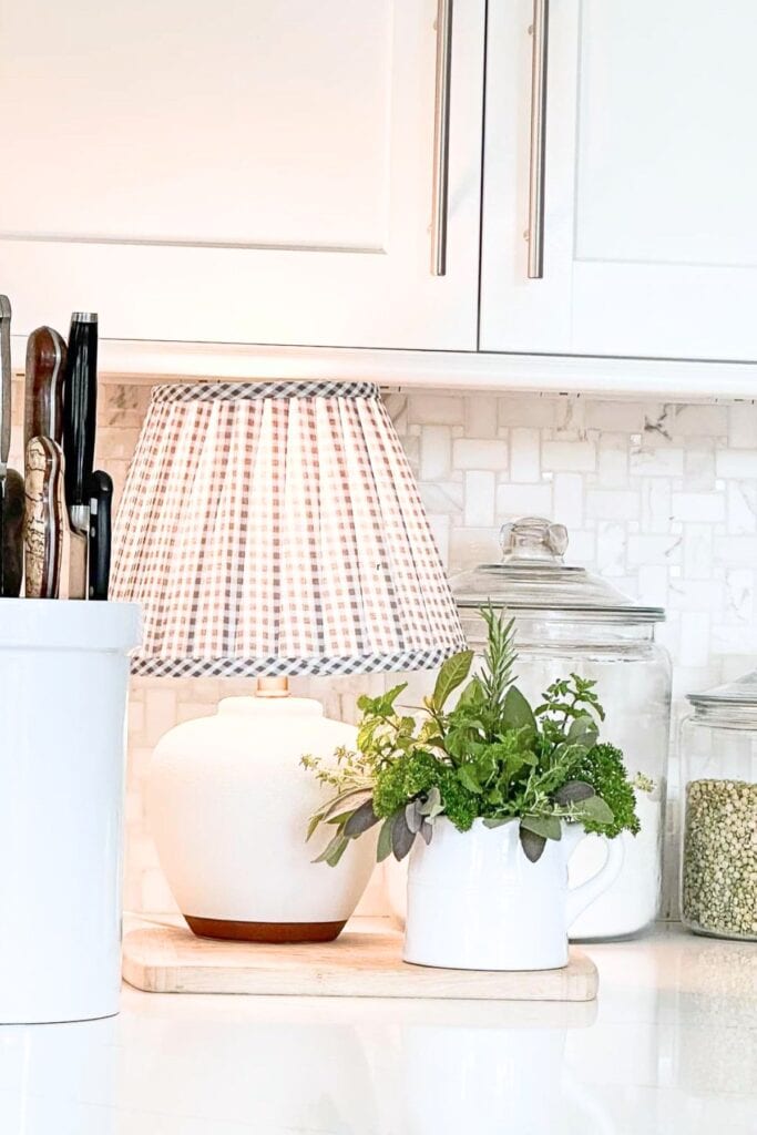 greenery arrangement in a white container on a kitchen counter next to a lamp and glass jars