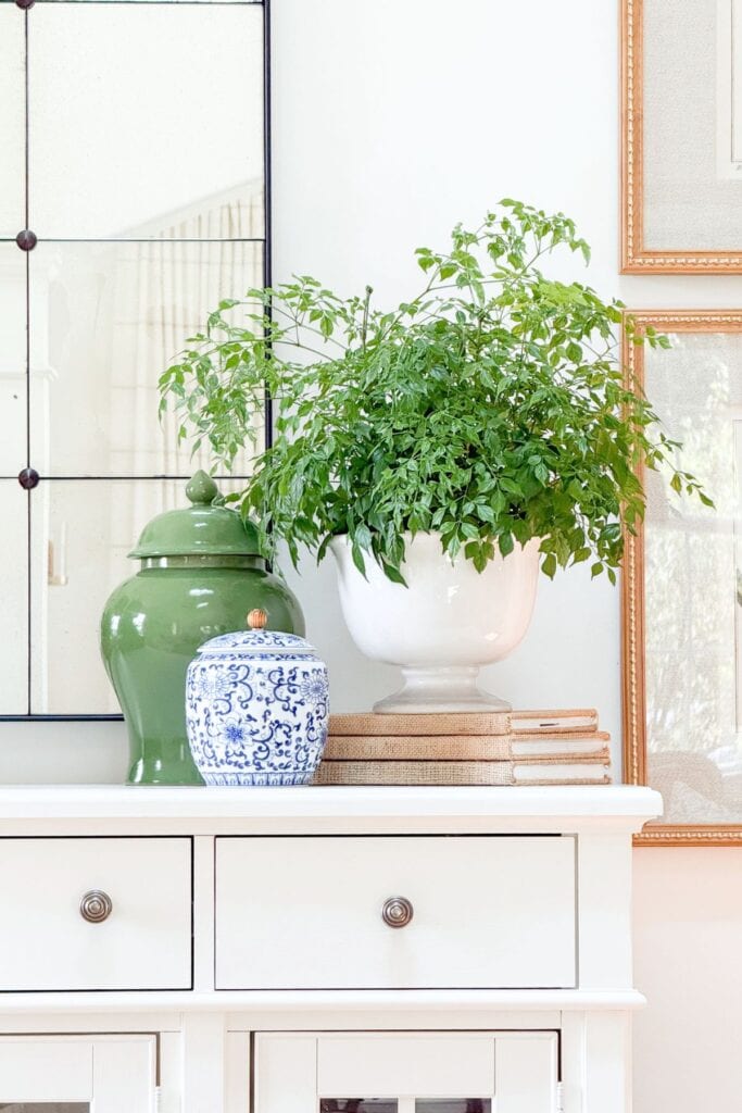 White buffet styled with a green ginger jar, blue and white jar, and greenery in a white urn atop burlap covered books