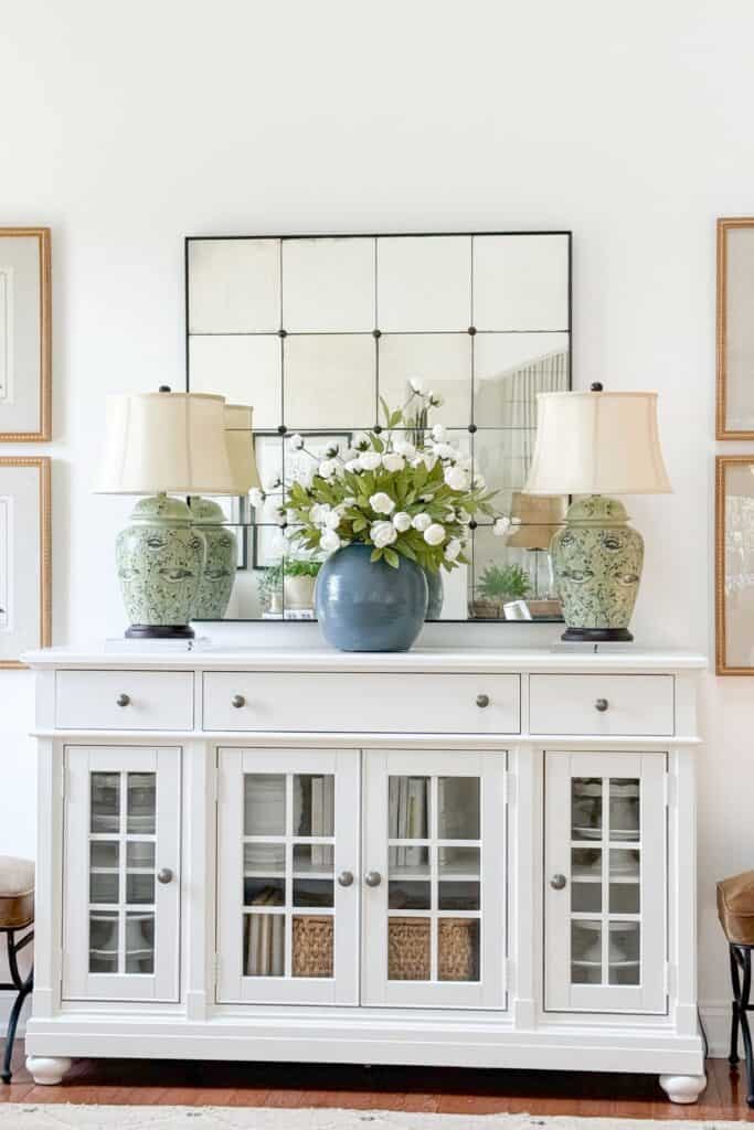 White buffet decorated with a pair of green chinoiserie lamps, an antique mirror, and a blue vase of white florals