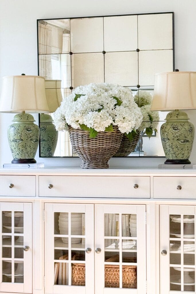neutral buffet styled with white hydrangeas in a basket, green lamps, and a mirror for balanced home decorating
