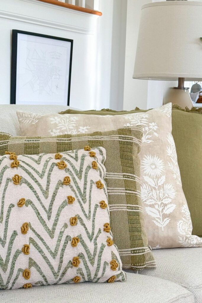 neutral sofa styled with layered pillows in green tones, blue and white vase, and greenery for cohesive decorating