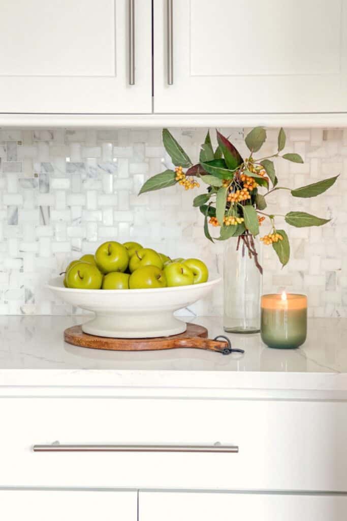 Kitchen countertop decor with a pedestal bowl of green apples, a berry branch, and a candle