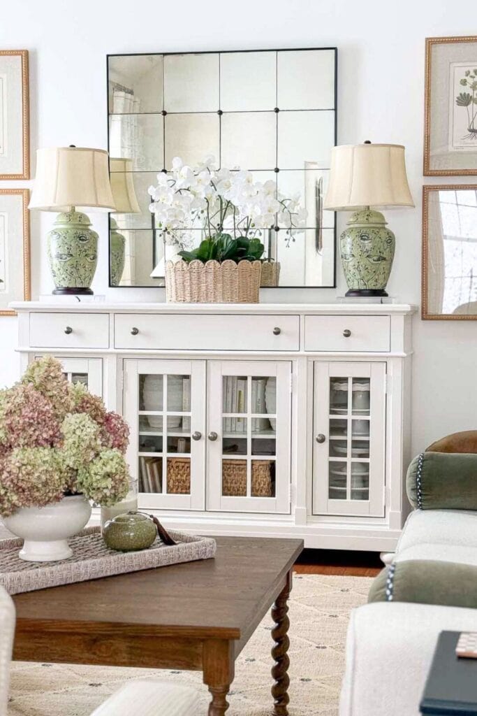 White buffet styled with green ceramic lamps, botanical artwork, a large mirror, and layered decor using balance and repetition.