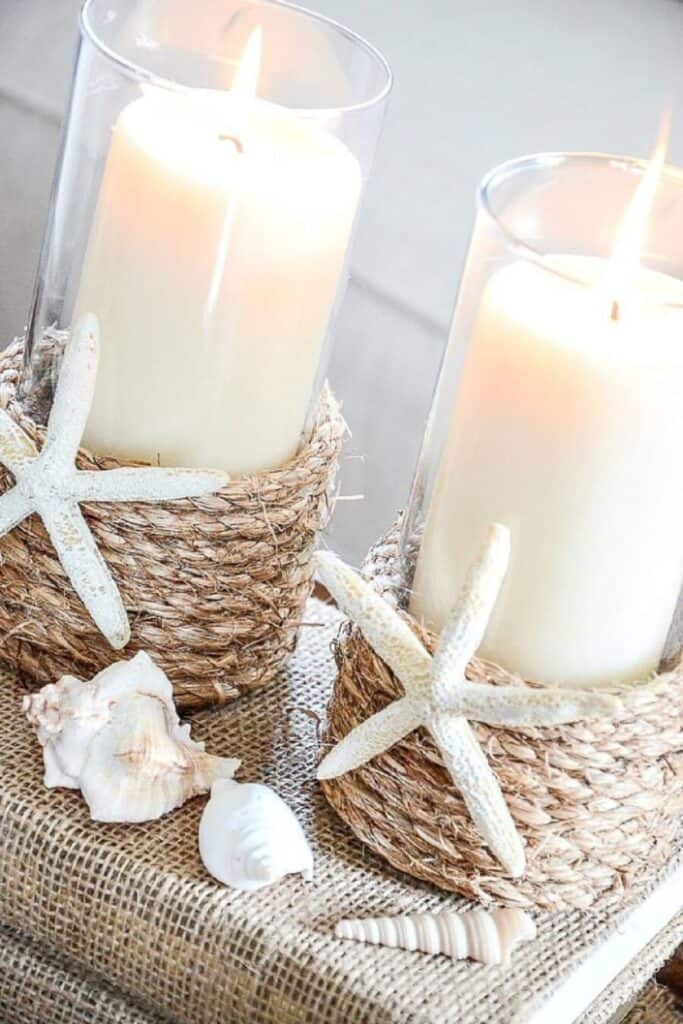 Two rope wrapped glass hurricane candle holders with white pillar candles, white starfish, and seashells arranged on a burlap and woven tray for a coastal summer vignette