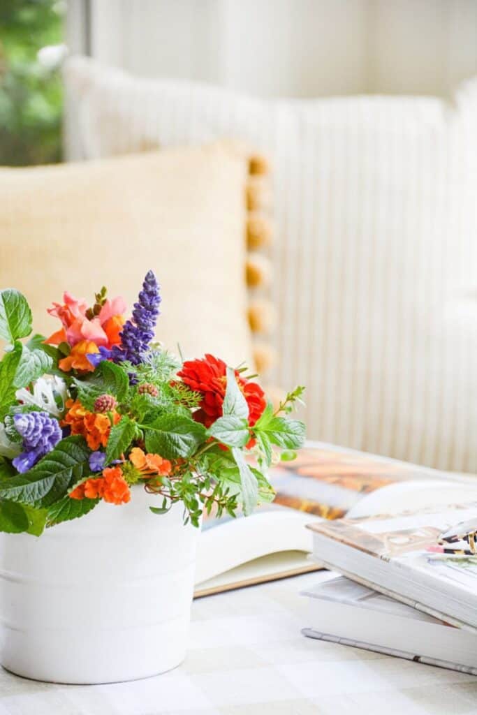 Colorful summer garden flower arrangement in a white cylindrical vase with red zinnias, purple salvia, orange lantana, pink snapdragons, and fresh greenery on a table with stacked magazines